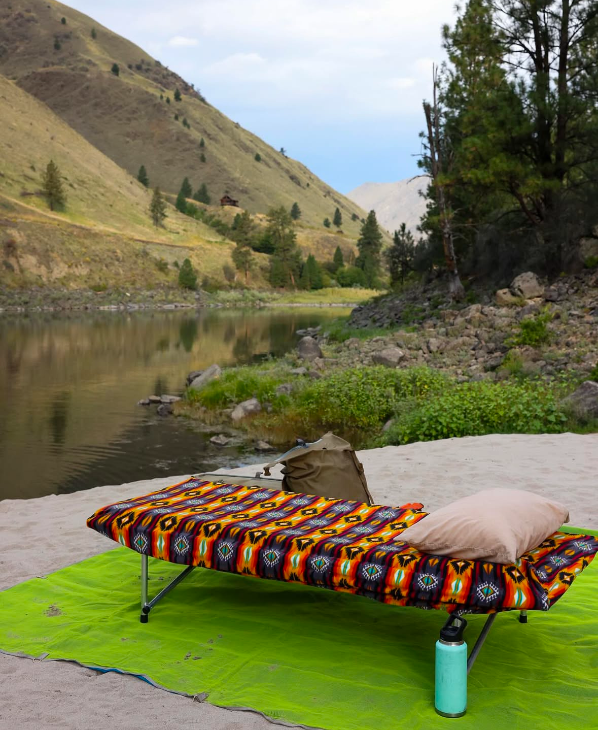 A vibrant FunLuvin' Cuddle Buddy from FunLuvin' Fleecewear, featuring a cozy fleece and cotton cover, is set up beside a river, surrounded by hills and trees. A backpack and water bottle are placed on the green mat beneath it, completing the ideal camping sleep gear setup.