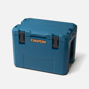 The "Outfitter V2 Series Coolers" by Canyon Coolers is a large blue portable cooler with a rectangular shape, sturdy latches, and premium soft-touch seal for excellent insulation. It features a Canyon logo and has a dual-action drain for easy emptying.