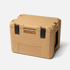 The tan, rugged Outfitter V2 Series Cooler by Canyon Coolers features black latches and a textured surface. It includes a dual-action drain and a premium soft-touch seal, ensuring your adventures stay cool.