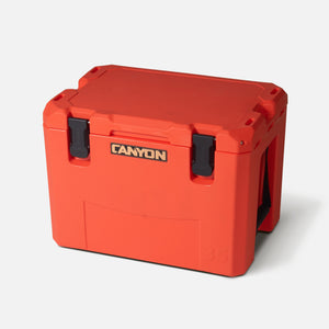 The Outfitter V2 Series Cooler by Canyon Coolers is a popular bright orange rectangular insulated cooler with flush square walls, secured by two black latches, featuring "CANYON" on the front and a dual-action drain for easy use.