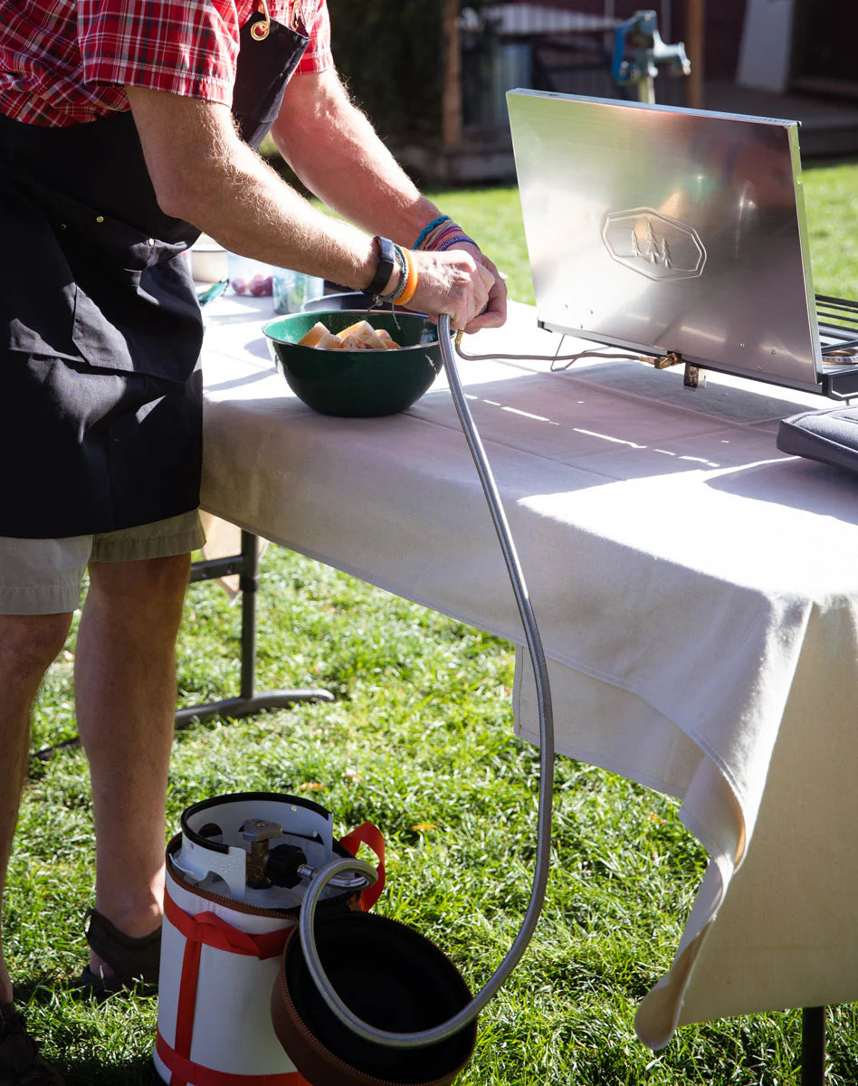 Person in a plaid shirt and apron setting up a portable gas stove outdoors, carefully making the camp stove connection with a GSI Propane Adapter Hose to a larger tank conversion.