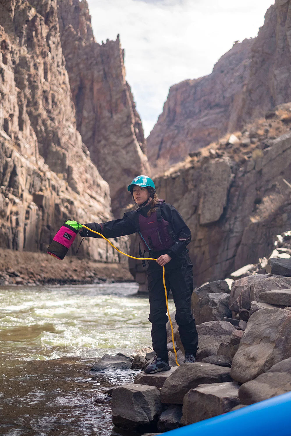 A person holding a River Station Gear B.O.A.T. - Classic Rescue Throw Bag - 70ft and a water container stands on rocks beside a river in a canyon.