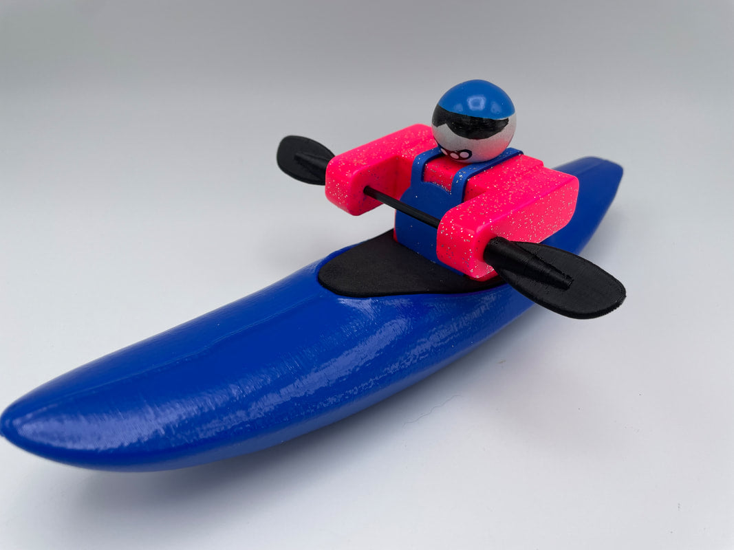 The Foamie Creeker by Foamie Friends features a figure kayaking in a vivid blue kayak with pink arms. Equipped with a stylish blue helmet and sunglasses, it's all set to tackle any waterway while gripping durable black paddles.