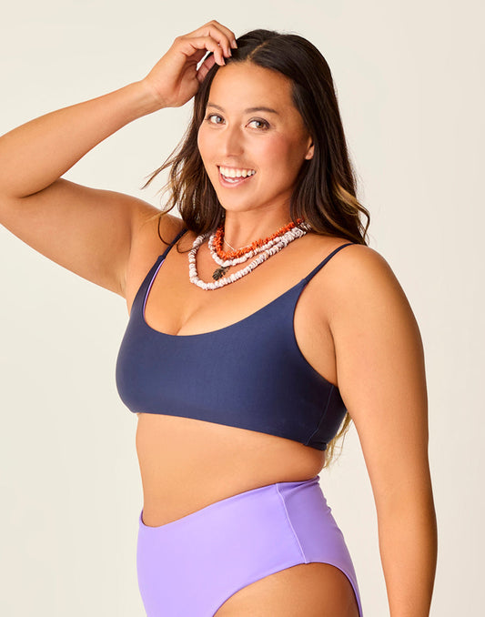 A person smiling, wearing a Carve Escondido Compression Reversible Top in navy blue and purple high-waisted bottoms, accessorized with stacked coral necklaces against a plain background.