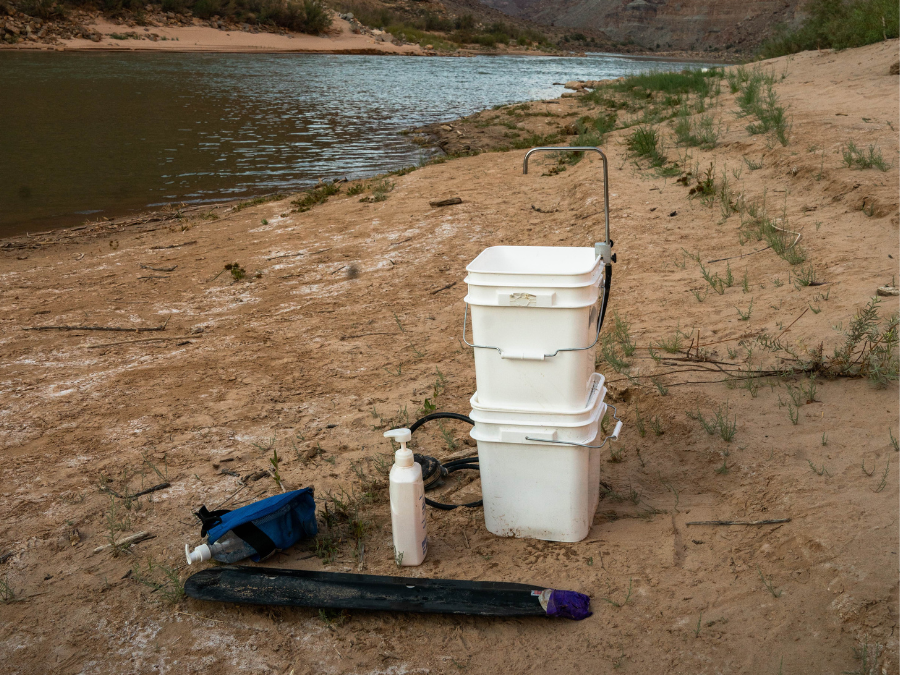 The Handwash Station by Down River Equipment is perfect for camping trips with two stacked white buckets, a blue bag, and a soap dispenser. A discreet black item accompanies them on the sandy riverbank near the water.