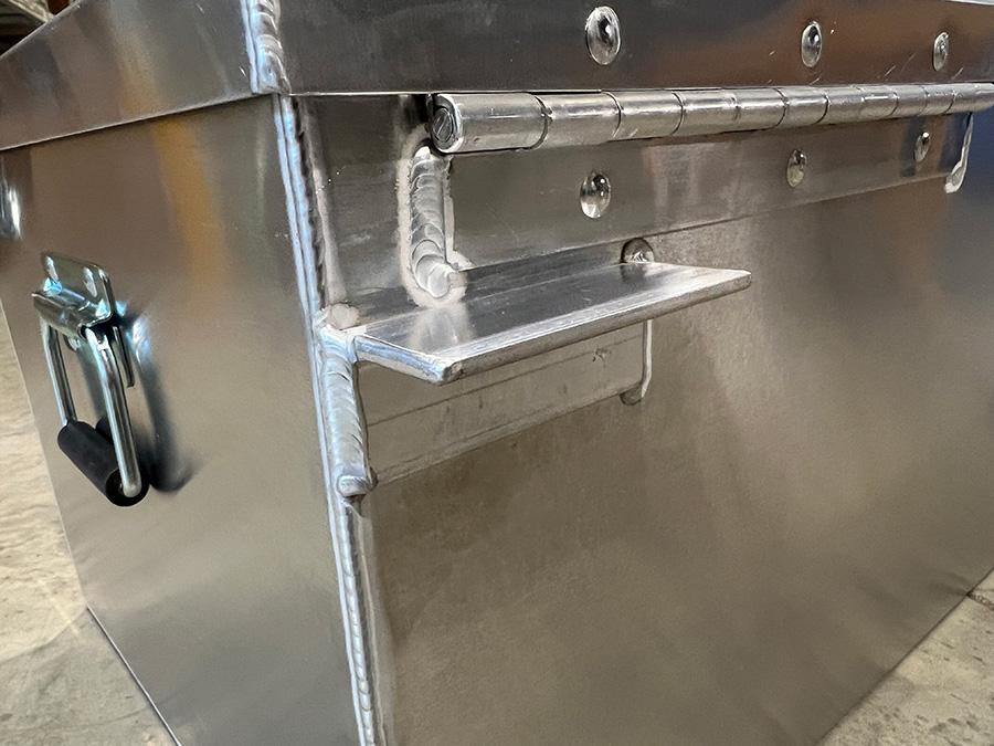 A close-up of the Down River Equipment Gunnison Drybox showcases its aircraft-grade aluminum construction, featuring a hinge and handle. Its sturdy build is highlighted by the welded seam and side rivets, while a rubber gasket ensures watertight security.