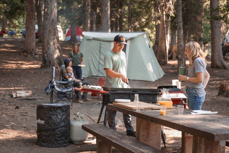 A group of people gathered around a picnic table in the woods, practicing Leave No Trace (LNT) principles with the Camp Chef Collapsible Garbage Can.