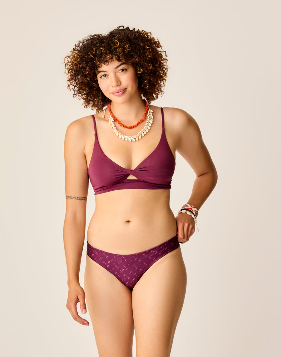 Wearing Carve's Sanitas 2.0 Compression Bottom in burgundy, a person with curly hair accessorizes with jewelry and stands against a plain backdrop.