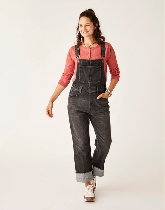 A woman in a Carve pink long-sleeve shirt and Jason Denim Overall stands against a plain backdrop, smiling and looking to the side. She is wearing white sneakers and has her hair tied up.