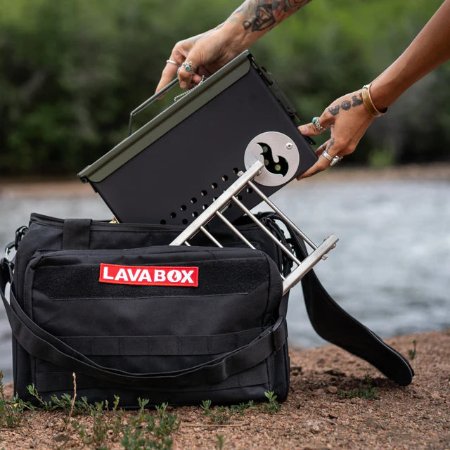 A person carefully places the Original LavaBox Combo firepit into a black bag labeled "LavaBox" beside the gentle river.