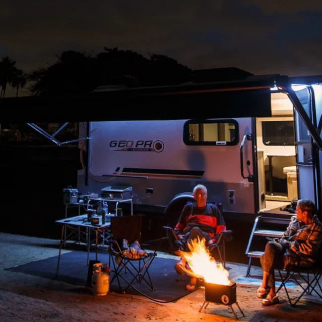 Under a camper van awning at night, two people sit next to the Original LavaBox Combo by LavaBox. A table with kitchen items is nearby, casting gentle shadows in the warm glow of the portable firepit.