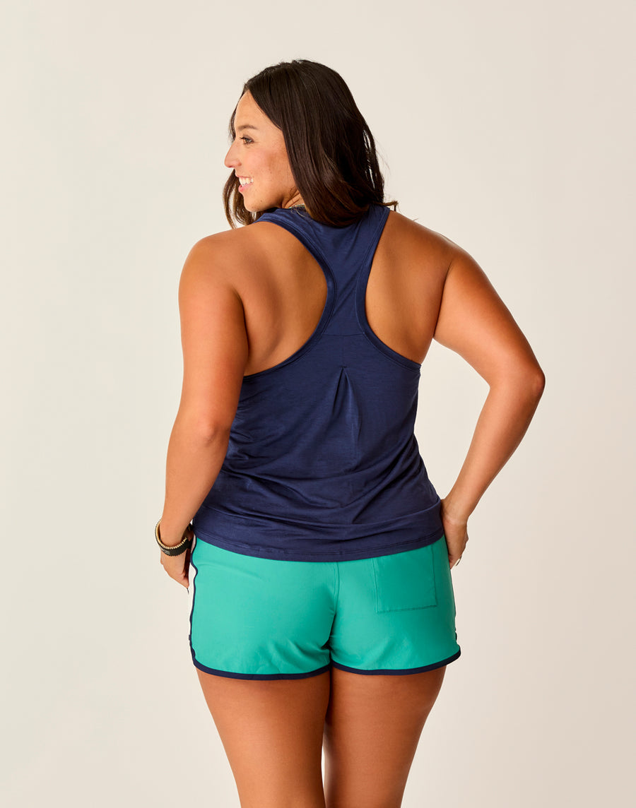 A person wearing a Tide Tank by Carve and green shorts stands smiling with one hand on their hip.