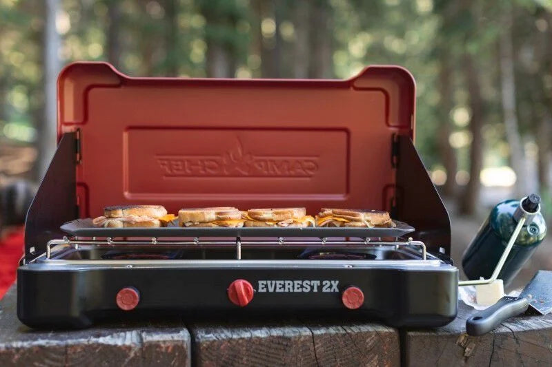 A red and black Camp Chef Mountain Series Steel Griddle is sitting on a table in the woods, perfect for outdoor cooking.