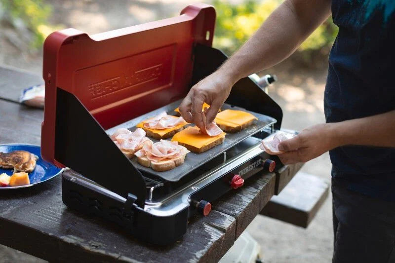 A man is preparing food on a Camp Chef Mountain Series Steel Griddle while outdoor cooking.