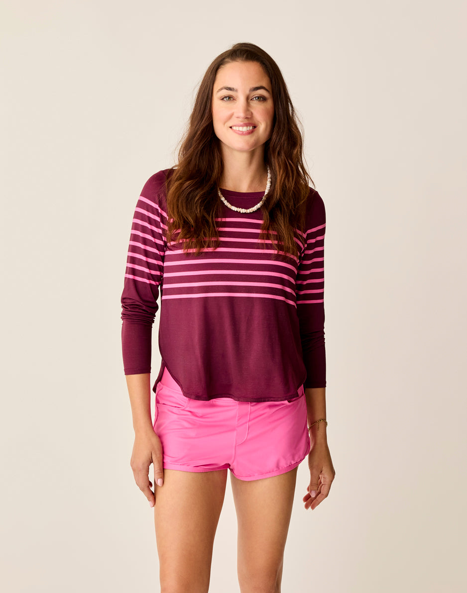 A woman with long hair smiles against a plain background, wearing Carve's Trek Top in maroon and pink stripes, matched with pink shorts and a necklace.