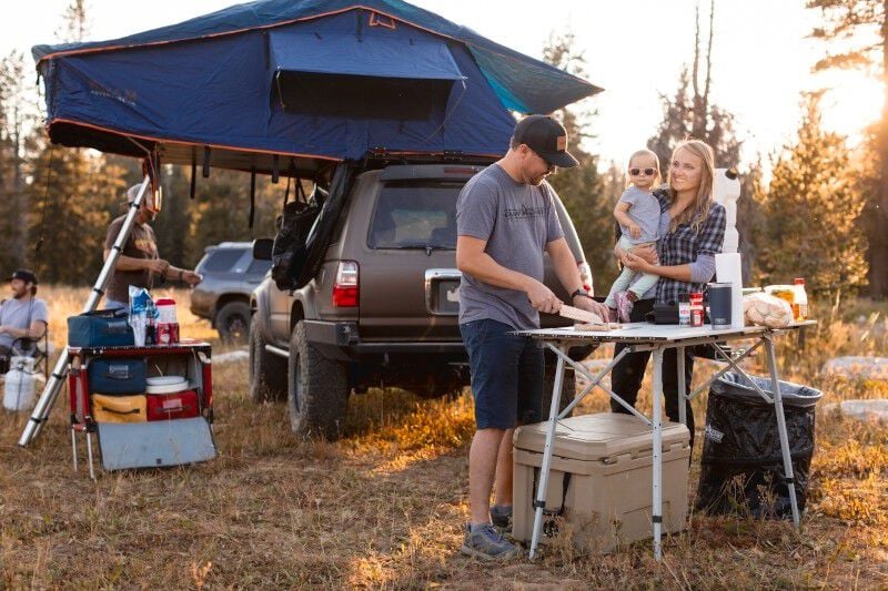 A group of people standing around a Camp Chef Mesa Adjustable Camp Table in the woods.