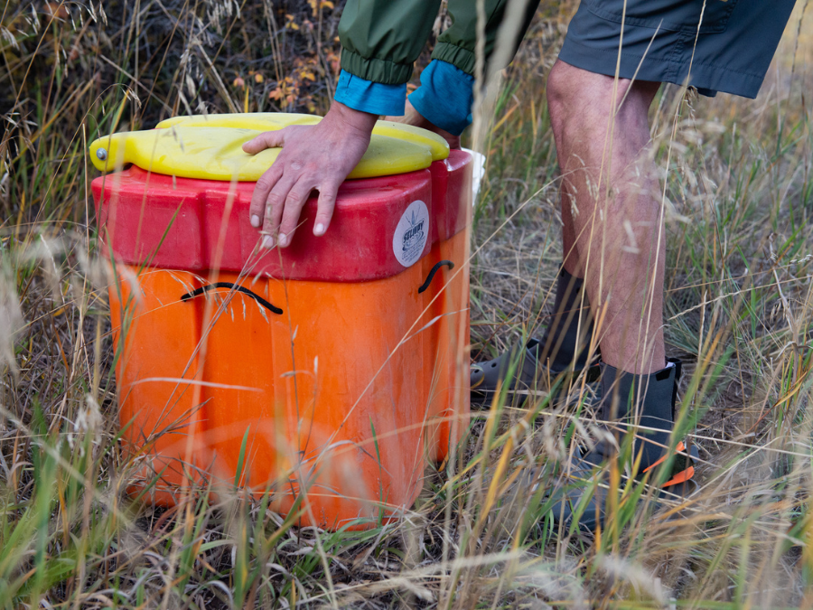 A person in boots secures a Selway River Bank II bear-proof container with a yellow lid by Down River Equipment outdoors in tall grass.