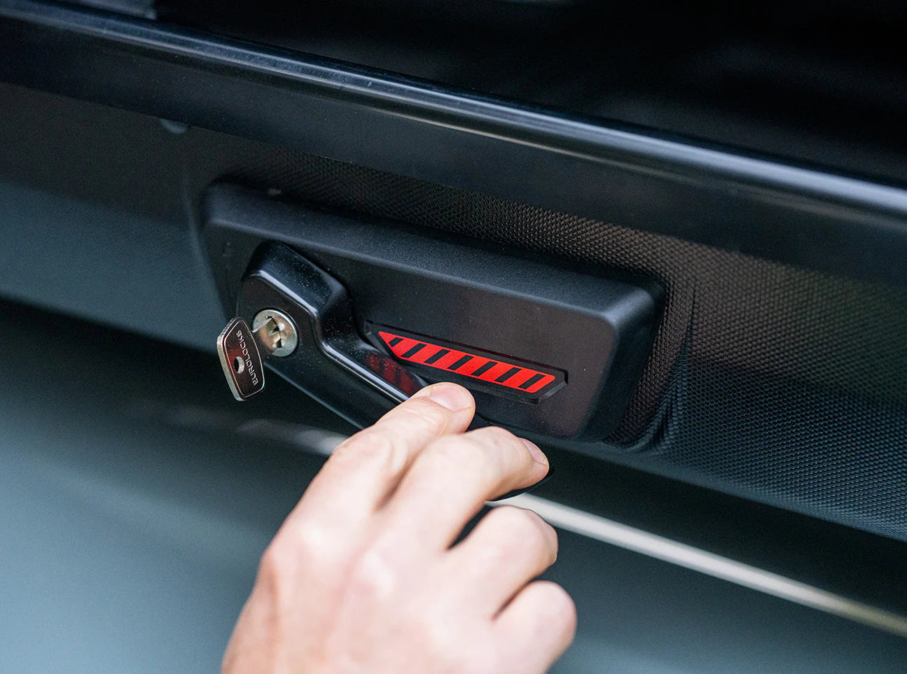 A hand unlocking a black compartment with a key reveals a red and black-striped label, akin to discovering the sleek storage potential of Yakima's SkyBox NX Skinny cargo box, all with tool-free installation ease.