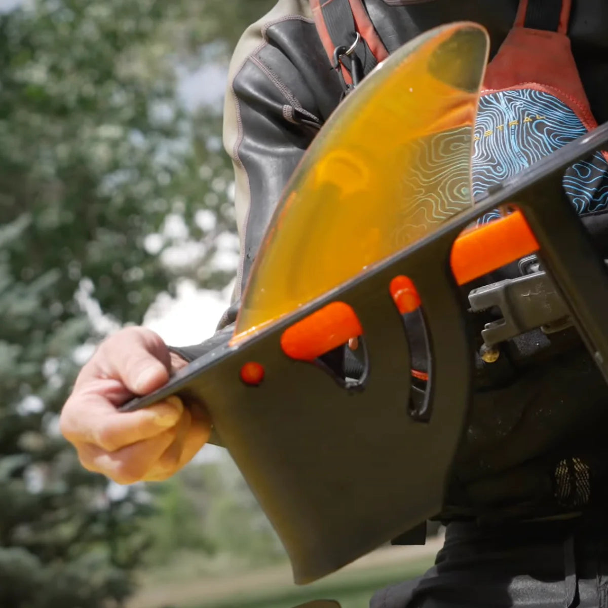 Among the trees, someone holds a black object with an elegantly curved, translucent orange fin, resembling the GalaXy SOLrivershine's sleek SUP design by SOL Paddle Boards. It features the Switchblade Retractable Fin System for seamless river adventures.