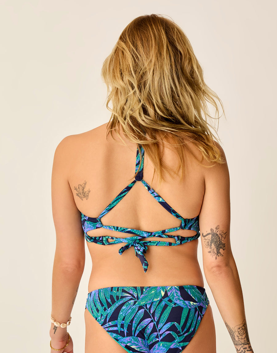 A person with long hair, wearing a blue and green leaf-patterned Carve Dahlia Top bikini, is seen from the back. The design features intricate straps and ties for a flattering fit, with visible arm tattoos beautifully accentuating the artistic active swimwear style.