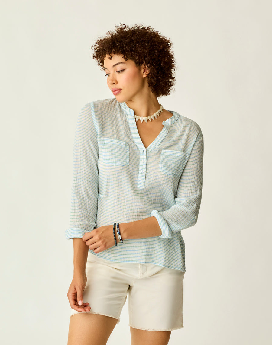 A woman with curly hair is posing against a plain background wearing a stylish Carve Dylan Gauze Shirt in light blue and pairing it with white shorts.